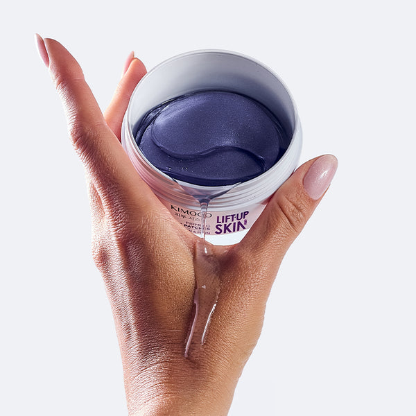Dłoń trzymająca otwarty pojemnik "Kimoco Lift-Up Skin Firming Hydrogel Eye Patches" z fioletowymi płatkami pod oczy, z płynem ściekającym po dłoni, na jasnym tle.