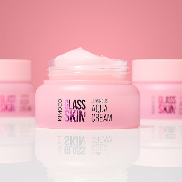 Zdjęcie na różowym tle: po środku otwarty różowy słoiczek "Kimoco Glass Skin Luminous Aqua Cream" z widocznym kremem na wierzchu, z rozmytymi słoiczkami w tle po obu stronach i odbiciem na lustrzanej powierzchni.
