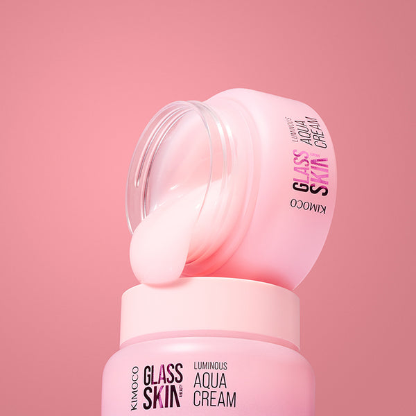 Zdjęcie na różowym tle: u góry przekrzywiona pokrywka słoiczka "Kimoco Glass Skin Luminous Aqua Cream" z kremem ściekającym na dolną część, u dołu stojący otwarty słoiczek tego samego produktu.