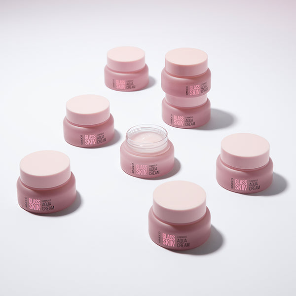 Zdjęcie na białym tle: po środku otwarty różowy słoiczek "Kimoco Glass Skin Luminous Aqua Cream" z widocznym kremem, otoczony siedmioma zamkniętymi słoiczkami tego samego produktu, ustawionymi w okręgu.