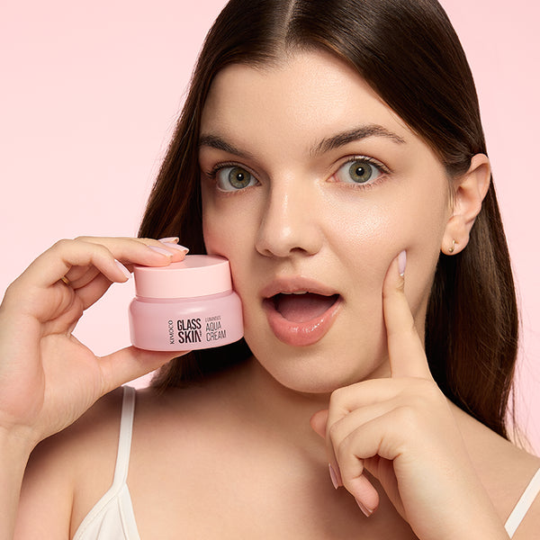 Zdjęcie na jasnoróżowym tle: z lewej kobieta trzymająca różowy słoiczek "Kimoco Glass Skin Luminous Aqua Cream" przy policzku, z prawej jej dłoń dotykająca drugiego policzka, twarz ze zdziwionym wyrazem i uchylonymi ustami.