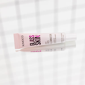 Po środku ułożona poziomo tuba "Kimoco Glass Skin Bright Eyes Illuminator" z widocznym odbiciem lustrzanym pod spodem, na jasnym tle z delikatnym wzorem kratki.