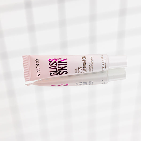 Po środku ułożona poziomo tuba "Kimoco Glass Skin Bright Eyes Illuminator" z widocznym odbiciem lustrzanym pod spodem, na jasnym tle z delikatnym wzorem kratki.
