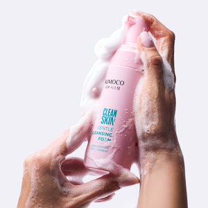 Dwie dłonie pokryte białą pianką trzymające różową butelkę z pompką Kimoco Clean Skin Gentle Cleansing Foam, na szarym tle.