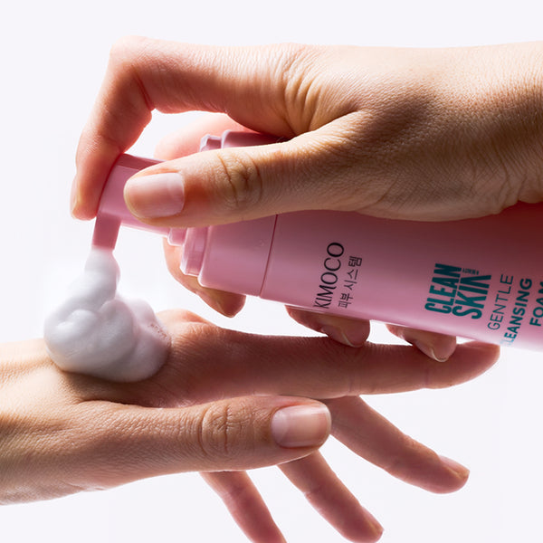 Dłoń trzymająca różową butelkę Kimoco Clean Skin Gentle Cleansing Foam, wyciskająca białą piankę na drugą dłoń z lewej strony, na białym tle.
