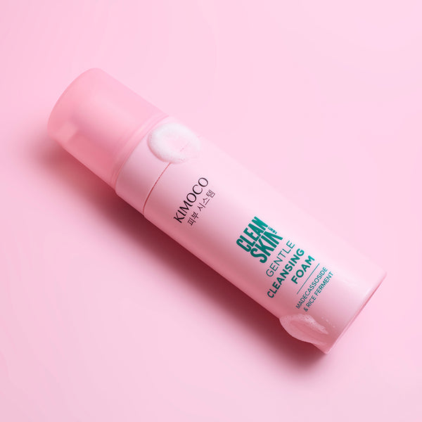 Po środku różowa butelka kosmetyku Kimoco Clean Skin Gentle Cleansing Foam z białą pianką przy pompce, leżąca ukośnie na różowym tle.