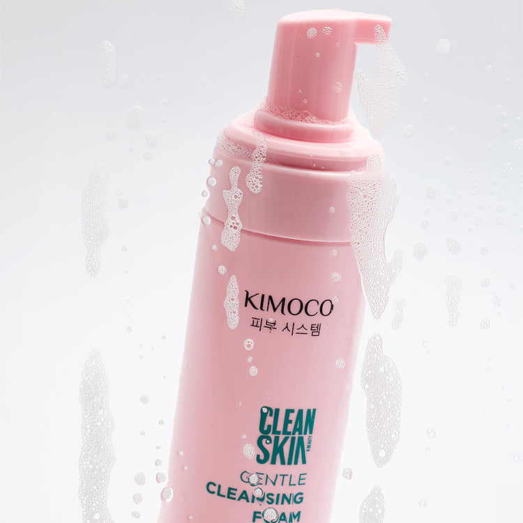 Po środku różowa butelka z pompką kosmetyku Kimoco Clean Skin Gentle Cleansing Foam, ustawiona ukośnie, otoczona z prawej strony bąbelkami pianki.