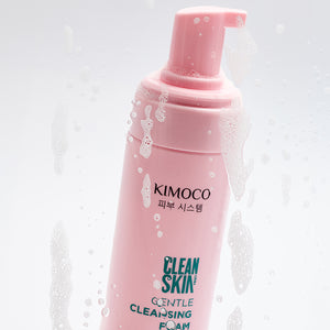Po środku różowa butelka z pompką kosmetyku Kimoco Clean Skin Gentle Cleansing Foam, ustawiona ukośnie, otoczona z prawej strony bąbelkami pianki.