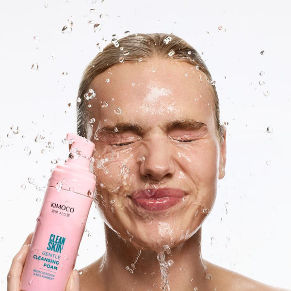Kobieta z blond włosami i zamkniętymi oczami, z wodą chlapiącą na twarz, trzymająca z lewej strony różową butelkę Kimoco Clean Skin Gentle Cleansing Foam, na białym tle.