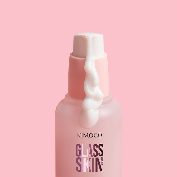 Zbliżenie na górną część różowej butelki kosmetyku Kimoco Glass Skin z pompką, z której spływa gęsta biała konsystencja produktu, na różowym tle.