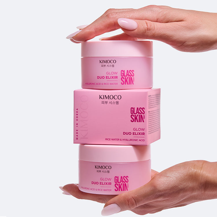 Zdjęcie na jasnoszarym tle: u góry dłoń dociskająca stos produktów "Kimoco Glass Skin Glow Duo Elixir" złożony z dwóch słoiczków i pudełka pomiędzy nimi, u dołu druga dłoń podtrzymująca stos od spodu.