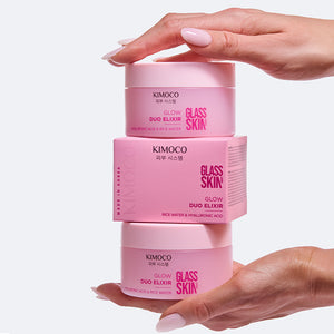 Zdjęcie na jasnoszarym tle: u góry dłoń dociskająca stos produktów "Kimoco Glass Skin Glow Duo Elixir" złożony z dwóch słoiczków i pudełka pomiędzy nimi, u dołu druga dłoń podtrzymująca stos od spodu.