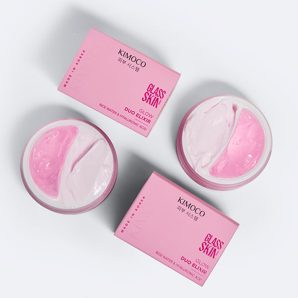 Zdjęcie na jasnoszarym tle: u góry i u dołu różowe pudełka "Kimoco Glass Skin Glow Duo Elixir", z lewej i z prawej otwarte słoiczki widziane z góry z dwoma masami — różową i białą.