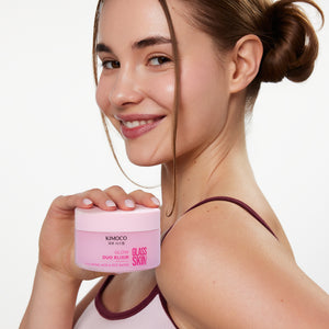Zdjęcie na białym tle: uśmiechnięta kobieta z włosami spiętymi w kok, trzymająca przy ramieniu różowy słoiczek "Kimoco Glass Skin Glow Duo Elixir".