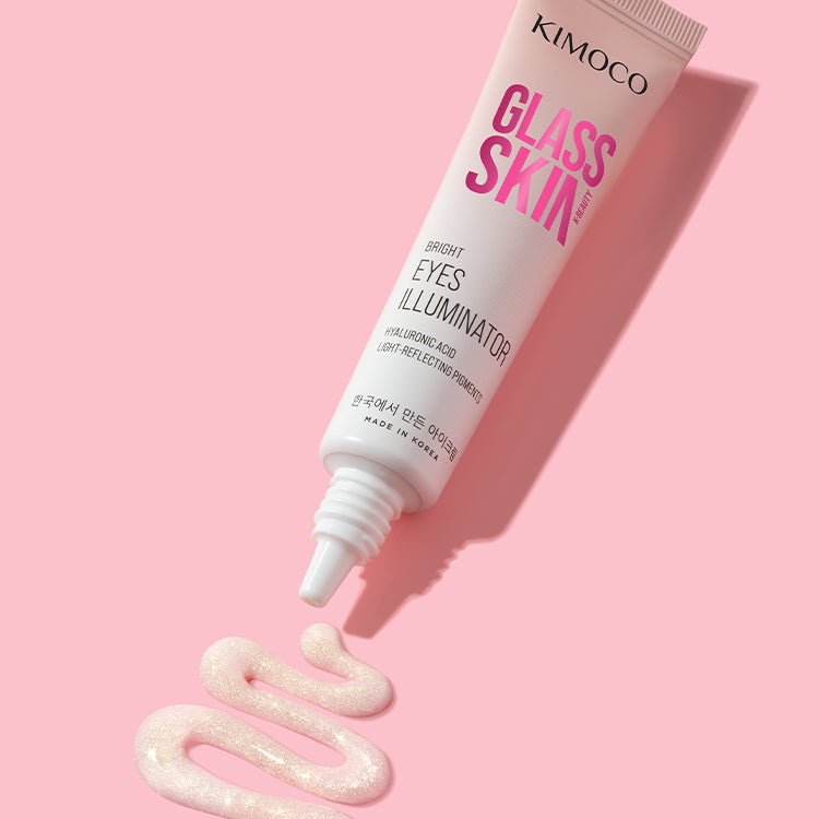 Zdjęcie na różowym tle: u góry ułożona ukośnie tuba "Kimoco Glass Skin Bright Eyes Illuminator", u dołu wyciśnięty z niej rozświetlający produkt w kształcie faliście ułożonej linii z widocznym brokatem.