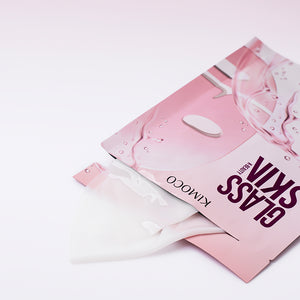 Opakowanie "Kimoco Glass Skin" z wysuwającą się z niego złożoną hydrożelową maseczką, na jasnym tle.