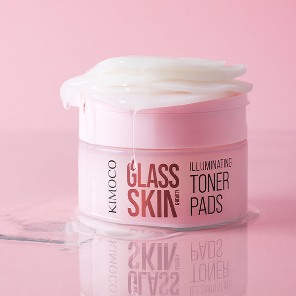 Pojemnik "Kimoco Glass Skin Illuminating Toner Pads" z płatkami wystającymi z wieczka i ściekającym płynem, z odbiciem na lustrzanej powierzchni, na różowym tle.