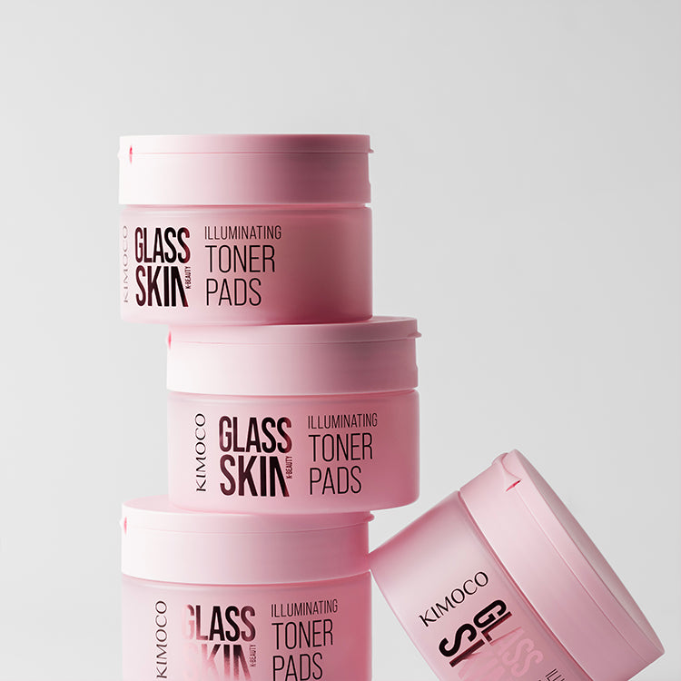 Pojemnik "Kimoco Glass Skin Illuminating Toner Pads" z płatkami wystającymi z wieczka i ściekającym płynem, z odbiciem na lustrzanej powierzchni, na różowym tle.