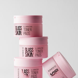 Ustawione w stos pojemniki "Kimoco Glass Skin Illuminating Toner Pads" z jednym pojemnikiem leżącym obok na boku, na jasnoszarym tle.