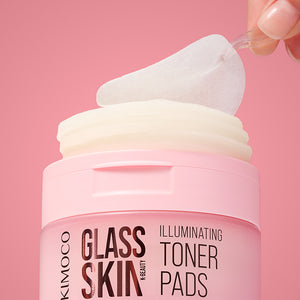 Plastikowa pęseta unosząca jeden płatek z otwartego pojemnika "Kimoco Glass Skin Illuminating Toner Pads" wypełnionego stosem płatków, na różowym tle.