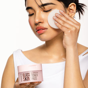 Kobieta z zamkniętymi oczami przecierająca policzek płatkiem, trzymająca w drugiej dłoni pojemnik "Kimoco Glass Skin Illuminating Toner Pads", na białym tle.