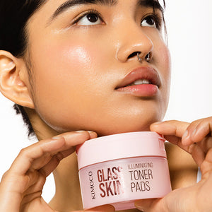 Kobieta patrząca w górę z dłońmi trzymającymi pod brodą pojemnik "Kimoco Glass Skin Illuminating Toner Pads", na białym tle.
