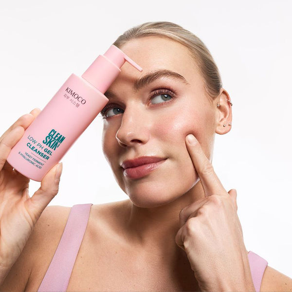 Kobieta z blond włosami spiętymi do tyłu, trzymająca z lewej strony różową butelkę z pompką kosmetyku Kimoco Clean Skin Low pH Gel Cleanser, dotykająca palcem prawego policzka.