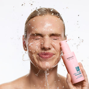 Kobieta z blond włosami i zamkniętymi oczami, z wodą chlapiącą na twarz, trzymająca z prawej strony różową butelkę Kimoco Clean Skin Gentle Cleansing Foam, na białym tle.