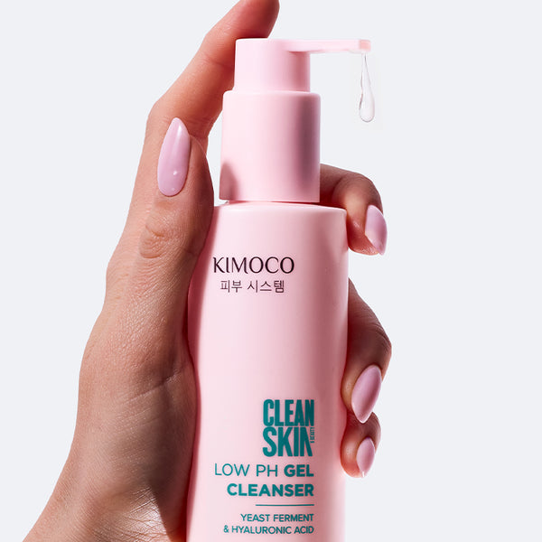 Dłoń z różowym manicure trzymająca różową butelkę z pompką kosmetyku Kimoco Clean Skin Low pH Gel Cleanser, z której kapie przezroczysty żel, na białym tle.