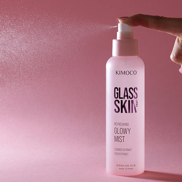 Palce naciskające pompkę różowej butelki kosmetyku Kimoco Glass Skin Refreshing Glowy Mist, z widoczną mgiełką produktu rozpylającą się w lewą stronę, na różowym tle