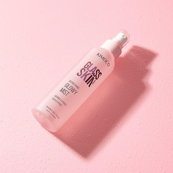 Różowa butelka z rozpylaczem kosmetyku Kimoco Glass Skin Refreshing Glowy Mist leżąca ukośnie, na różowym tle pokrytym kropelkami wody