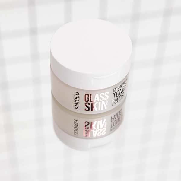 Zamknięty pojemnik "Kimoco Glass Skin Moisturizing Toner Pads" z odbiciem lustrzanym pod spodem, na jasnym tle z delikatnym wzorem kratki.