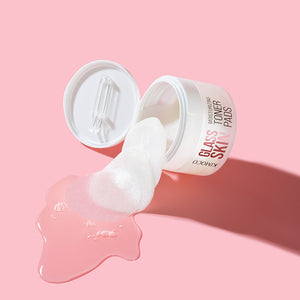 Przewrócony na bok pojemnik "Kimoco Glass Skin Moisturizing Toner Pads" z wysuwającymi się płatkami i wylanym płynem, obok odłożona pokrywka z plastikowymi cążkami, na różowym tle.