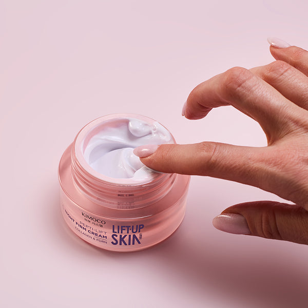 Z lewej strony otwarty różowy słoiczek kosmetyku Kimoco Lift-Up Skin z białym kremem, z prawej strony dłoń nabierająca krem palcem wskazującym, na różowym tle