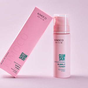 Po środku różowe pudełko oparte o różową butelkę z pompką kosmetyku Clean Skin Smoothing Bubble Toner, na fioletowo-różowym tle.