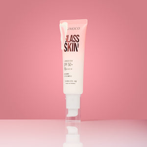 Zdjęcie na różowym tle: po środku stojąca tuba "Kimoco Glass Skin Luminous Dew SPF 50+ PA++++" z odkręconą przezroczystą zakrętką, z odrobiną kremu wypływającego z góry tuby, z odbiciem na lustrzanej powierzchni.