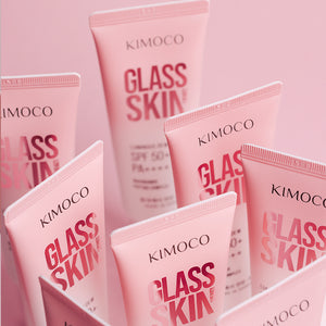Zdjęcie na różowym tle: po środku wyższa tuba "Kimoco Glass Skin" ustawiona pionowo z zakrętką na górze, otoczona z lewej i z prawej mniejszymi tubami tego samego produktu ustawionymi obok siebie.