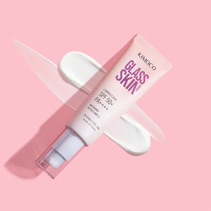 Zdjęcie na różowym tle: po środku ułożona ukośnie tuba "Kimoco Glass Skin Luminous Dew SPF 50+ PA++++" z odkręconą zakrętką, w tle rozsmarowany biały krem po przekątnej.