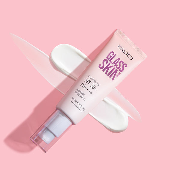 Zdjęcie na różowym tle: po środku ułożona ukośnie tuba "Kimoco Glass Skin Luminous Dew SPF 50+ PA++++" z odkręconą zakrętką, w tle rozsmarowany biały krem po przekątnej.