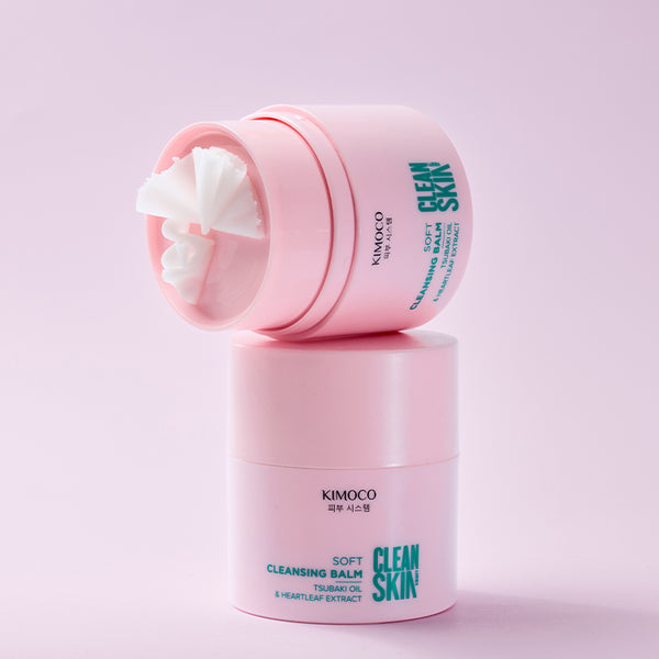 Po środku otwarty słoiczek kosmetyku Kimoco Clean Skin Soft Cleansing Balm z białym balsamem w środku, oparty na drugim zamknietym słoiczku tego samego produktu, na różowym tle.