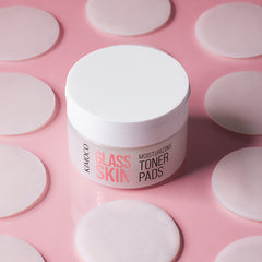 Zamknięty pojemnik "Kimoco Glass Skin Moisturizing Toner Pads" otoczony rozłożonymi okrągłymi płatkami, na różowym tle.
