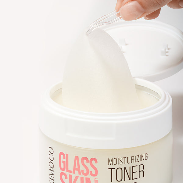 Plastikowa pęseta unosząca płatek z otwartego pojemnika "Kimoco Glass Skin Moisturizing Toner Pads", z odłożoną obok pokrywką, na jasnym tle.