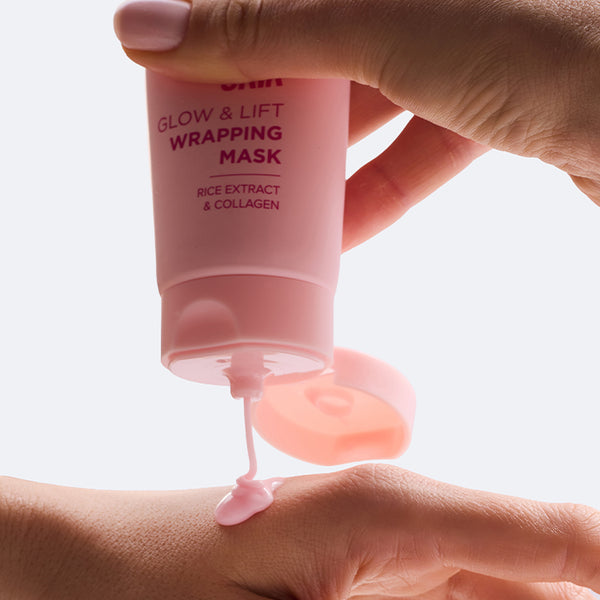 Dłoń trzymająca różową tubkę Glow & Lift Wrapping Mask z ekstraktem ryżowym i kolagenem, z otwartej tubki wypływa produkt skapujący na dłoń drugiej ręki znajdującą się poniżej, na jasnym tle.
